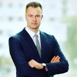 Portret mężczyzny w granatowym garniturze z założonymi rękami, widoczny zegarek na lewym nadgarstku, jasne tło biurowe.