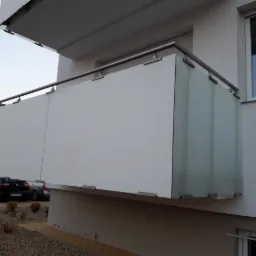 Balkon z nowoczesną balustradą, łączącą białe panele z elementami ze stali nierdzewnej i szkła matowego, widok z dołu na tle parkingu i budynku mieszkalnego.