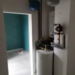 Biały zasobnik ciepłej wody użytkowej Bosch z podłączonymi rurami i osprzętem na tle białej ściany, widoczne również manometry, w tle ściana z turkusową tapetą z motywem roślinnym.