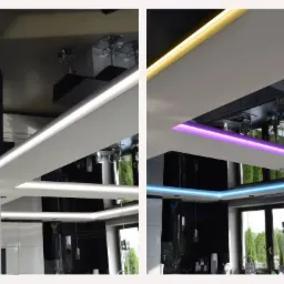 Nowoczesny sufit podwieszany z oświetleniem LED w trzech kolorach: białym, fioletowym i żółtym, odbijający się w błyszczącej czarnej powierzchni, widoczne okno i zieleń na zewnątrz.