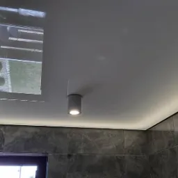 Nowoczesna łazienka z szarą marmurową okładziną ścienną, podwieszanym sufitem z oświetleniem LED i punktową lampą, odbijającym krajobraz na zewnątrz.