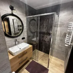 Nowoczesna łazienka z szarymi płytkami imitującymi kamień, umywalką nablatową z wzorem marmuru na drewnianej szafce, okrągłym lustrem w czarnej ramie i kabiną prysznicową ze ścianką z efektem rdzy.