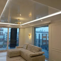 Nowoczesny salon z podwieszanym, lustrzanym sufitem z oświetleniem LED, widoczne złote lampy i narożnik w jasnym odcieniu.