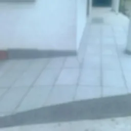 Wyłożone szarymi płytkami wejście do domu z rampą, widoczne fragmenty elewacji i kolumny podtrzymującej zadaszenie.