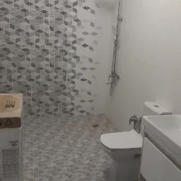 Wykończona łazienka w Olsztynie z szarymi, geometrycznymi płytkami na podłodze i ścianie, kabina prysznicowa typu deszczownica oraz biała umywalka z szafką, widoczny karton z boku.