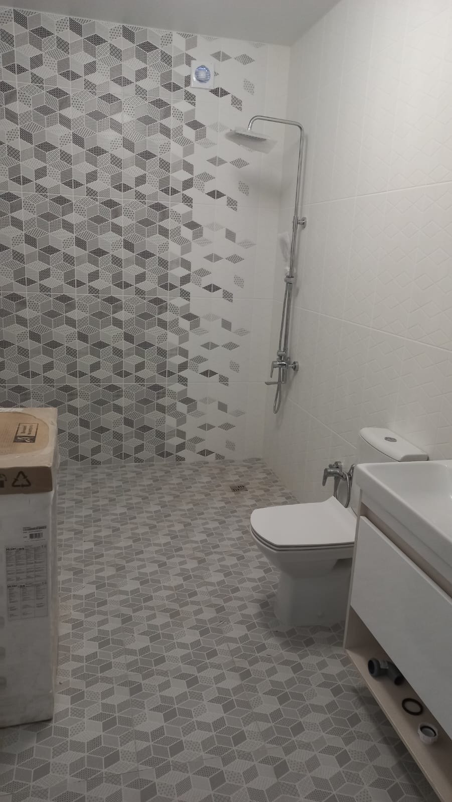 Wykończona łazienka w Olsztynie z szarymi, geometrycznymi płytkami na podłodze i ścianie, kabina prysznicowa typu deszczownica oraz biała umywalka z szafką, widoczny karton z boku.