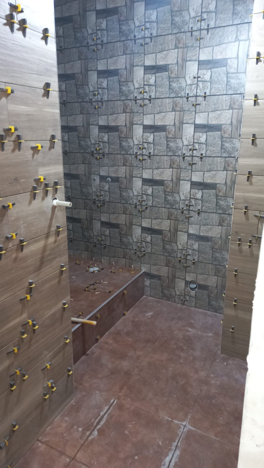 Układanie płytek w łazience, widoczne brązowe płytki podłogowe i imitujące kamień płytki ścienne z żółtymi krzyżykami dystansowymi, etap prac przed fugowaniem.