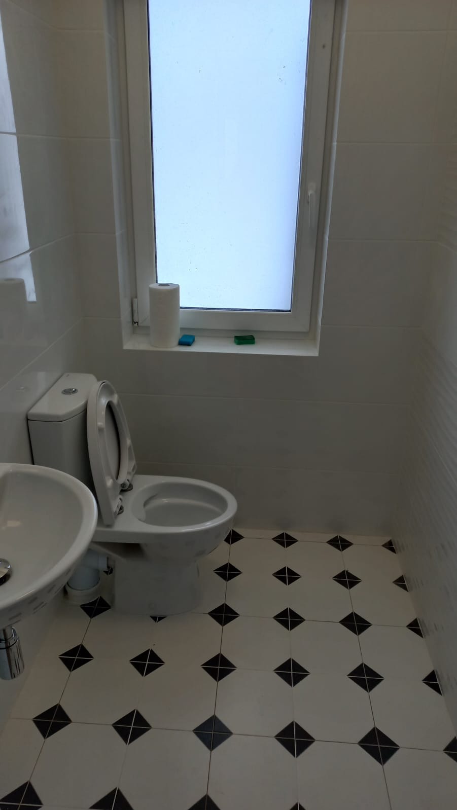 Wykończona łazienka z białą armaturą, płytkami podłogowymi w czarno-białe romby i oknem z parapetem, na którym stoją rolka papieru toaletowego i dwa małe mydełka.