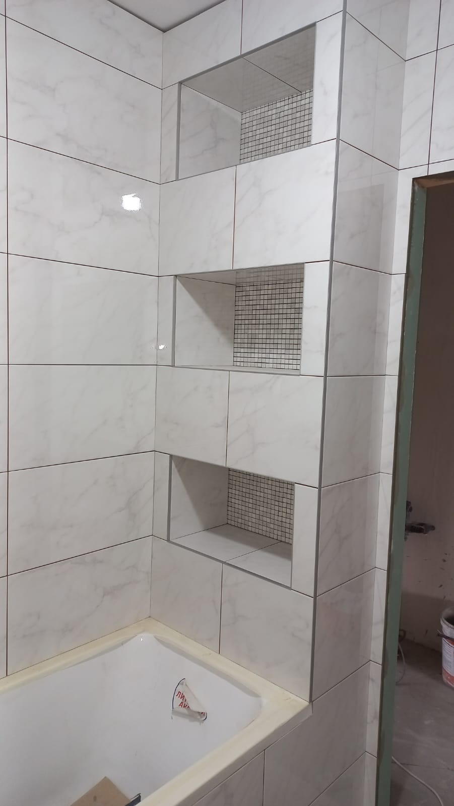 Wykończona łazienka z białymi marmurkowymi płytkami, wnęki z siatką drucianą, wanna z plastikową osłoną, widok z perspektywy narożnika.