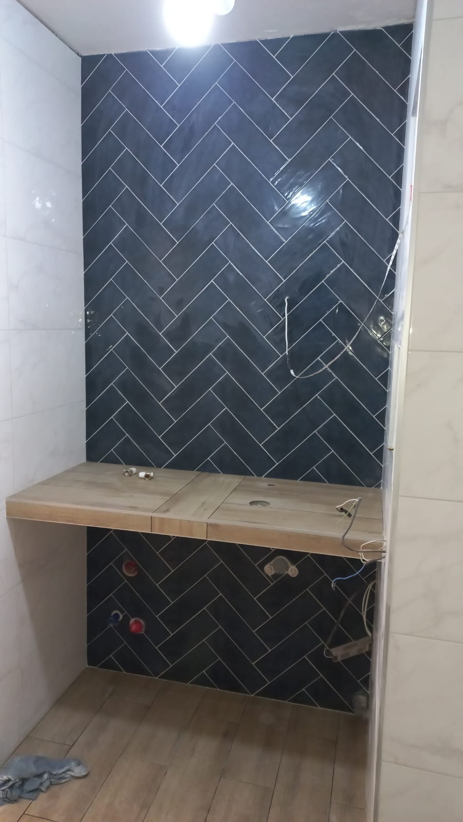 Montaż ciemnych płytek ułożonych w jodełkę na ścianie i pod blatem umywalkowym w łazience, widoczne przyłącza wodne i elementy instalacji elektrycznej.