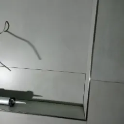 Wnętrze łazienki z szarymi płytkami, widoczny aplikator silikonu na półce i fragment instalacji elektrycznej.