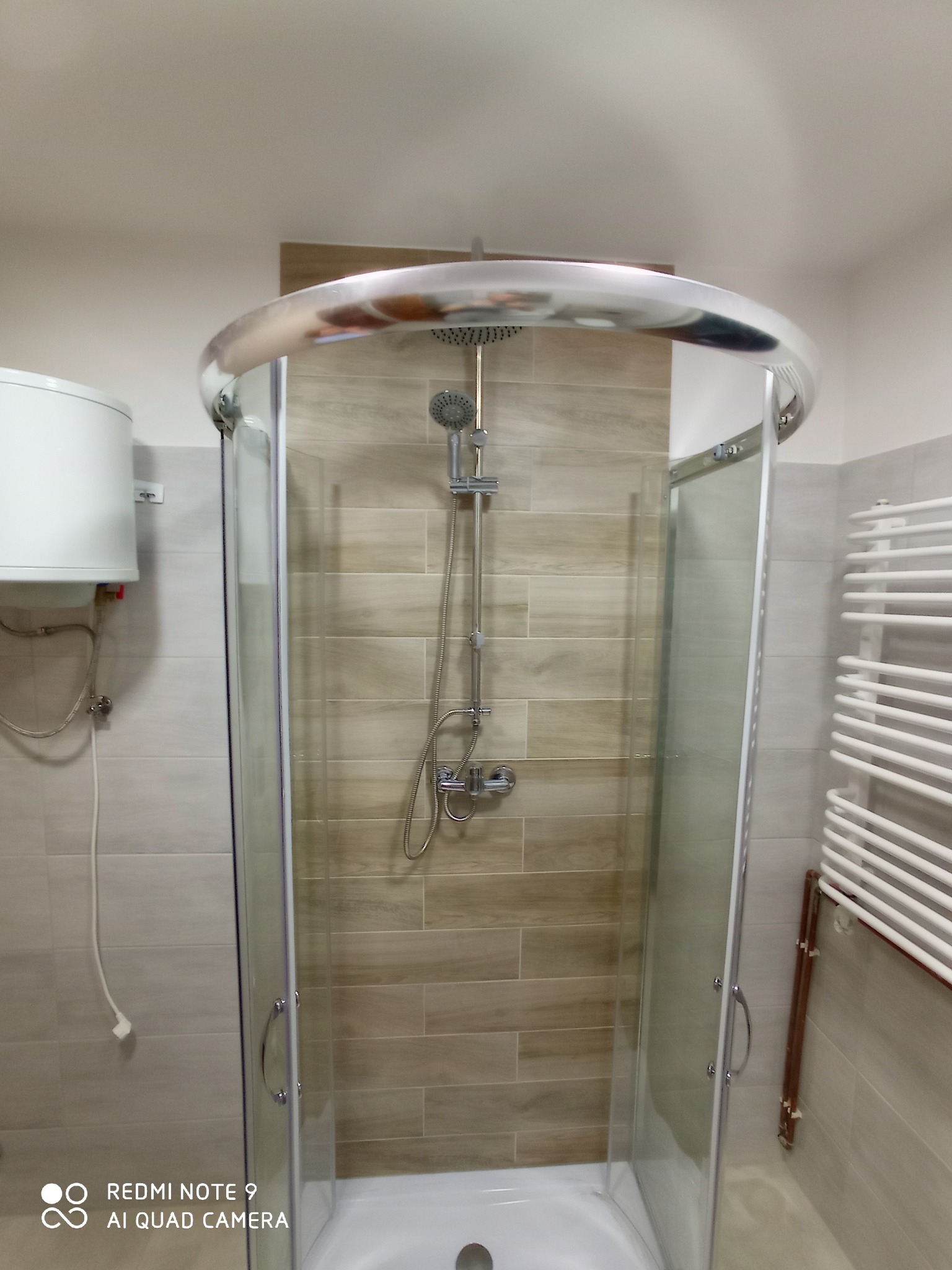 Kabina prysznicowa z zaokrąglonymi drzwiami i deszczownicą, na ścianie płytki imitujące drewno, obok grzejnik drabinkowy i bojler.