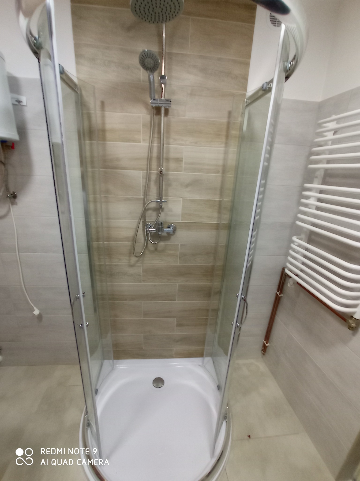 Kabina prysznicowa z chromowaną armaturą i deszczownicą, na ścianach beżowe płytki imitujące drewno, biały grzejnik drabinkowy po prawej stronie.