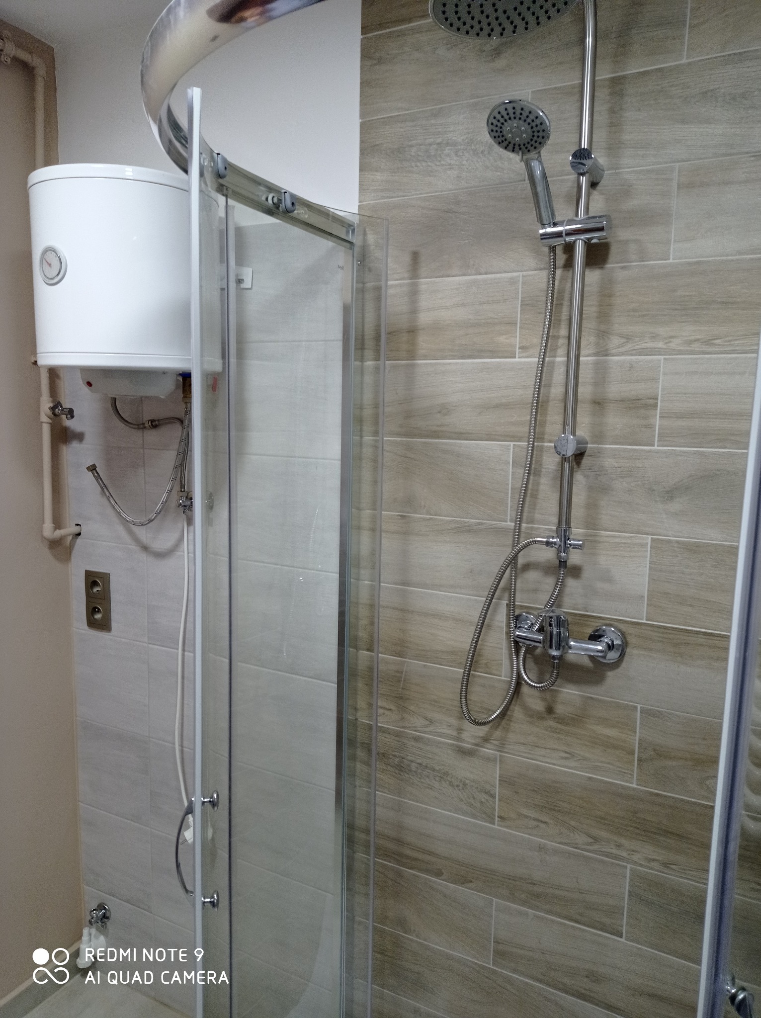 Wykończona łazienka z beżowymi płytkami imitującymi drewno, kabiną prysznicową z przesuwnymi drzwiami, bojlerem elektrycznym i zestawem prysznicowym z deszczownicą i słuchawką.