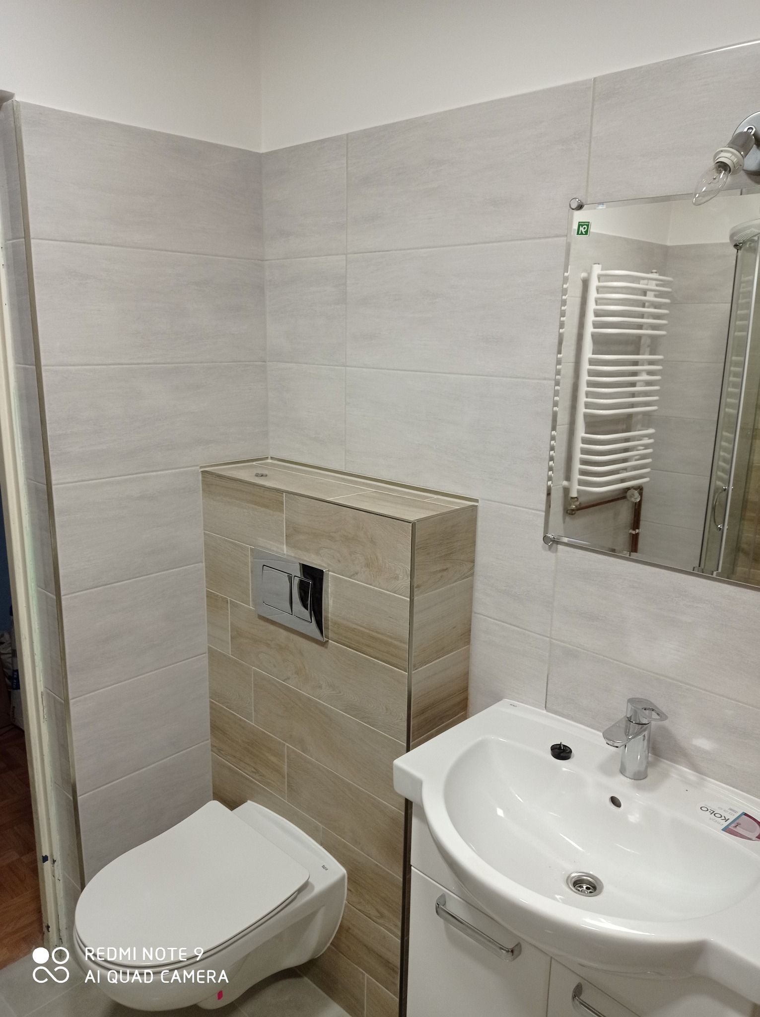 Nowoczesna łazienka z podwieszaną toaletą, umywalką z szafką i lustrem, wykończona płytkami w odcieniach szarości i beżu.