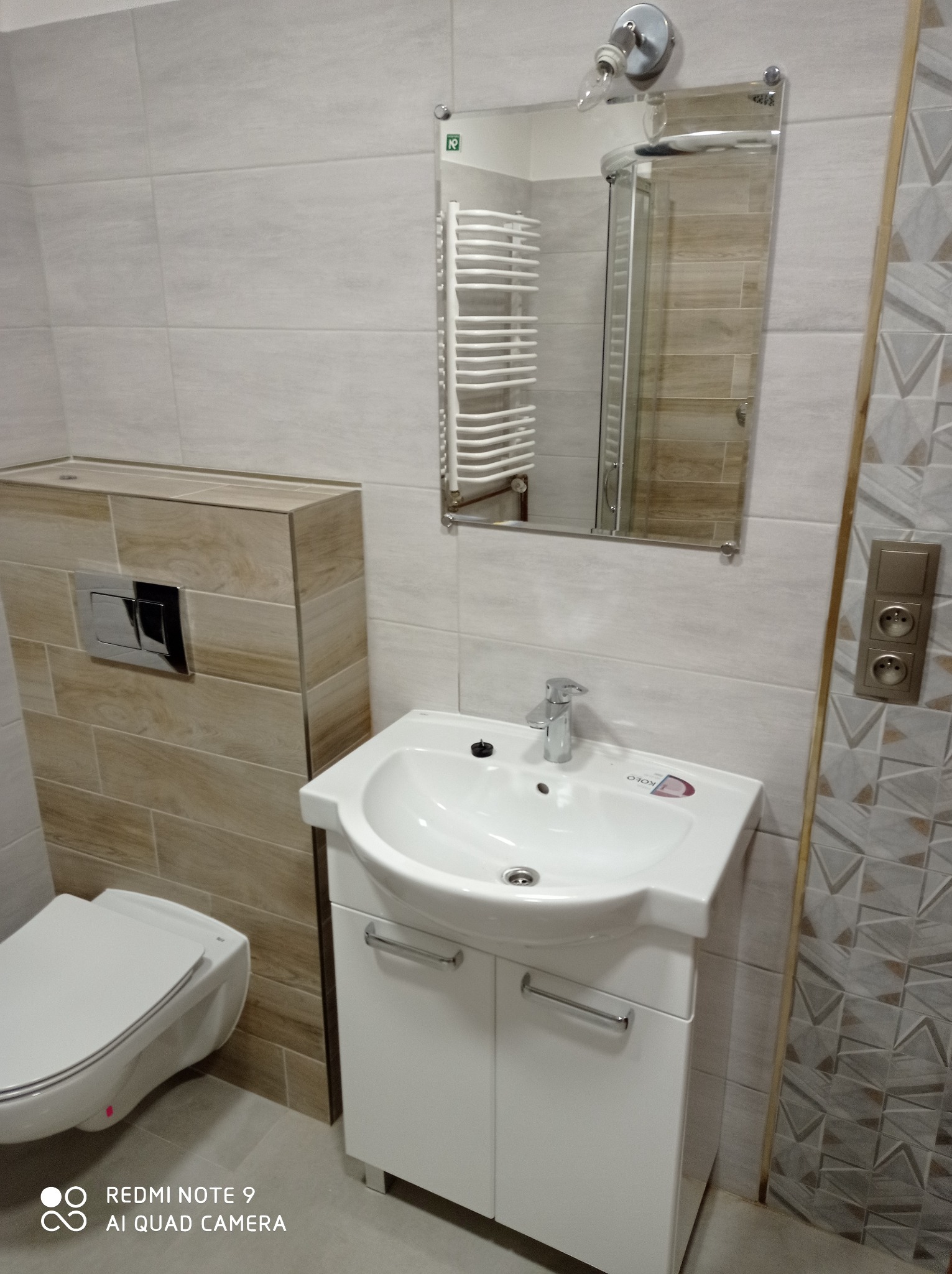 Ukończona toaleta z podwieszaną muszlą, umywalką z szafką i lustrem, wykończona płytkami w odcieniach beżu i szarości, widoczny fragment grzejnika drabinkowego w odbiciu lustra.