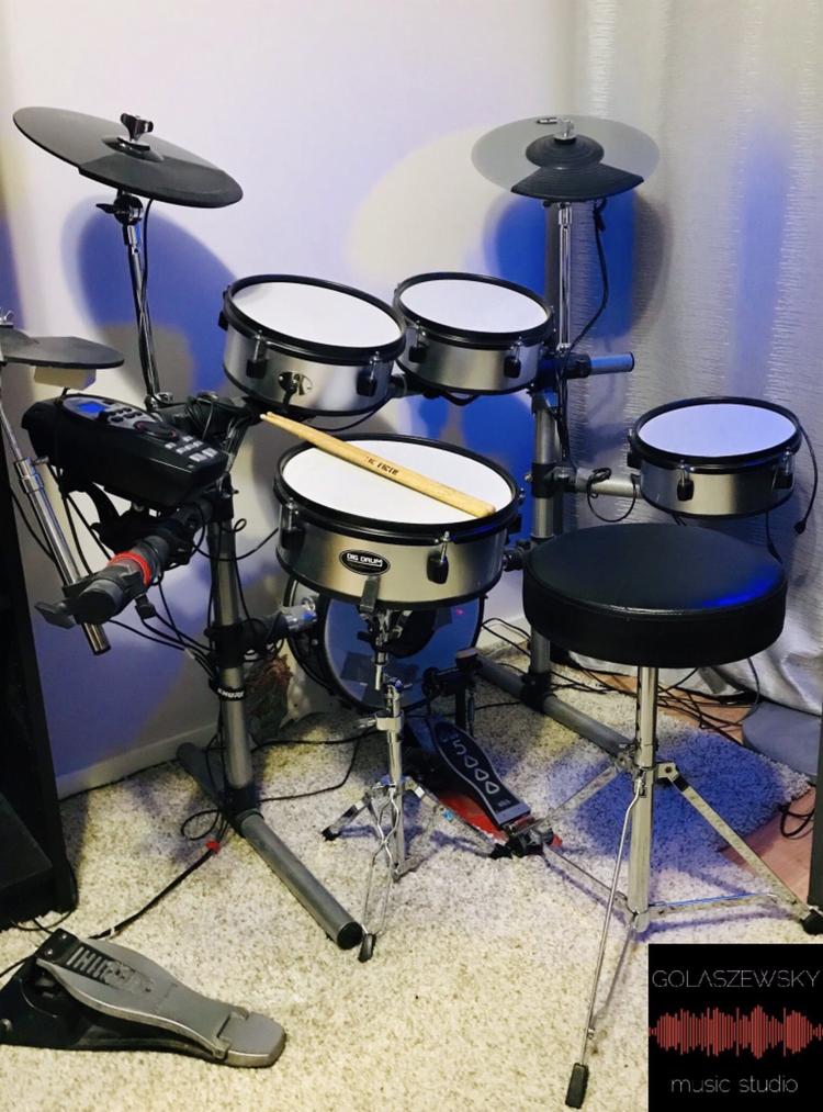 Elektroniczna perkusja z pałeczką na werblu, widoczny stołek perkusyjny i pedały, na podłodze dywan z długim włosiem, w rogu logo studia muzycznego.