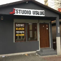 Fasada szarego budynku usługowego z szyldem 'STUDIO USŁUG' i czerwoną ikoną buta, okno z napisem 'SZEWC KLUCZE PIECZĄTKI OSTRZENIE', brązowe drzwi wejściowe i kosz na śmieci BIO z prawej strony.