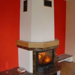 Klasyczny kominek obudowany marmurem w odcieniach brązu i bieli, z płomieniami widocznymi za szybą, na tle czerwonej ściany. Na półce kominka leżą dwie książki.