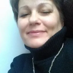 Portret uśmiechniętej kobiety w czarnym golfie i naszyjniku z pereł na tle jasnej ściany.