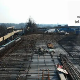 Zbrojenie stropu budynku w trakcie budowy, widok z góry na siatkę z prętów stalowych i drewniane szalunki, w tle zabudowa miejska.