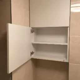 Biała szafka wisząca z otwartymi drzwiczkami w łazience z beżowymi płytkami, widoczne zawiasy i półki wewnątrz.