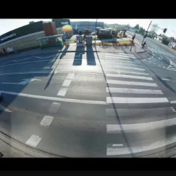 Szeroki kadr z kamery monitoringu ulicznego, ukazujący skrzyżowanie z przejściem dla pieszych, budynki i przejeżdżający samochód w słoneczny dzień.