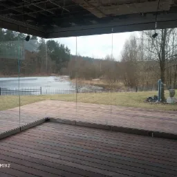 Szklana ściana tarasu z widokiem na częściowo zamarznięte jezioro i las w pochmurny dzień. Drewniana podłoga tarasu z desek w odcieniach brązu.