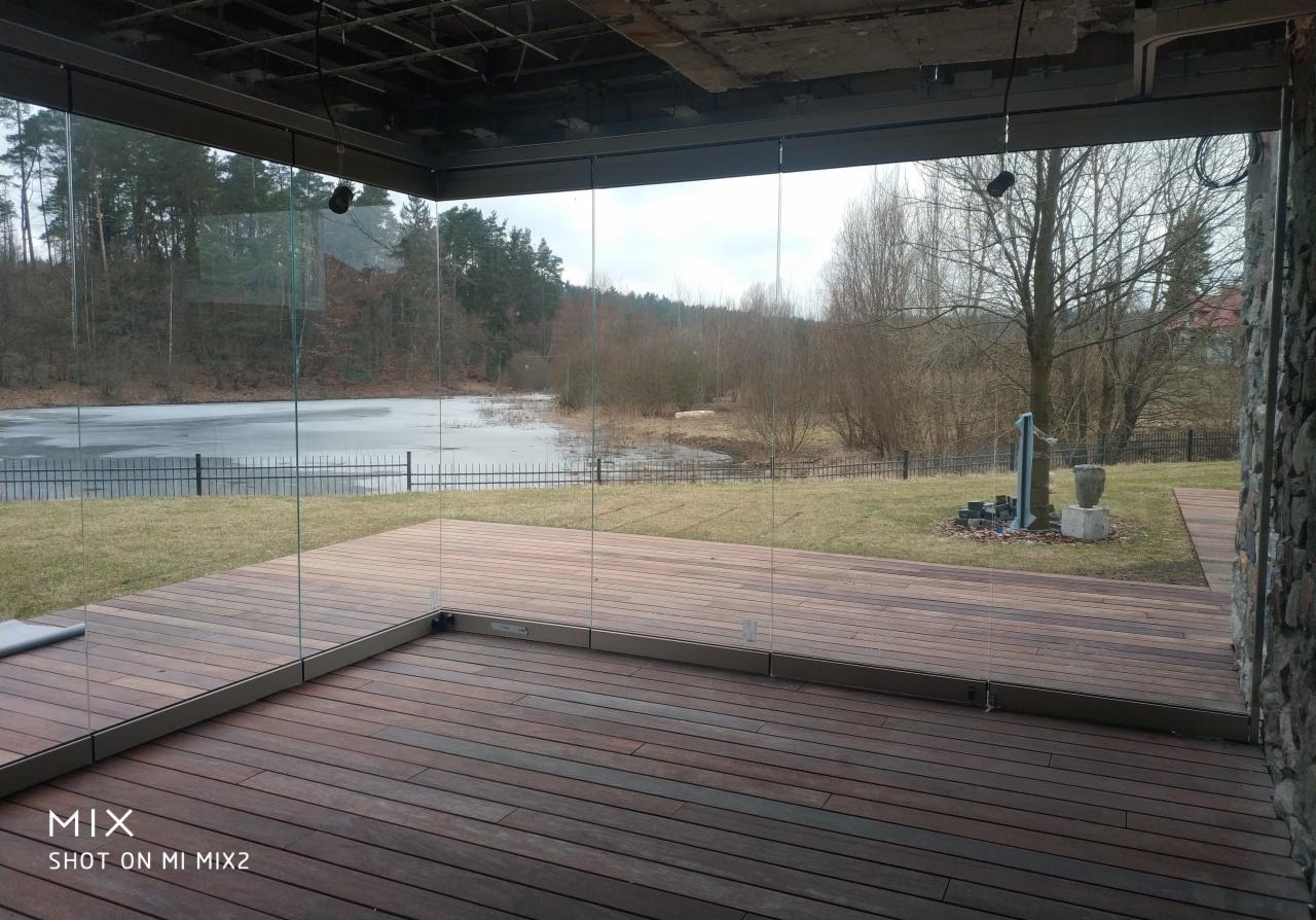 Szklana ściana tarasu z widokiem na częściowo zamarznięte jezioro i las w pochmurny dzień. Drewniana podłoga tarasu z desek w odcieniach brązu.