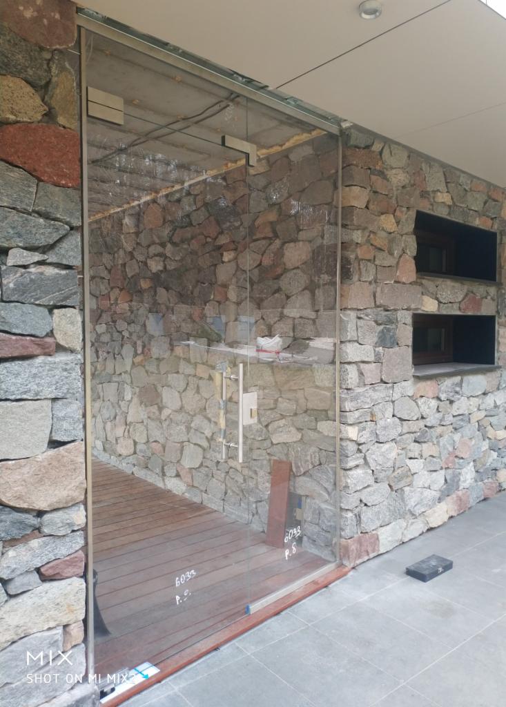 Szklane drzwi wejściowe z metalową ramą i podłużnym uchwytem, wbudowane w ścianę z nieregularnego kamienia, prowadzące na drewniany taras.