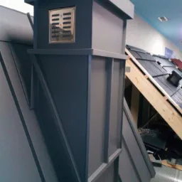 Szary komin wentylacyjny na dachu z blachy trapezowej, widoczne próbki dachówek w tle.