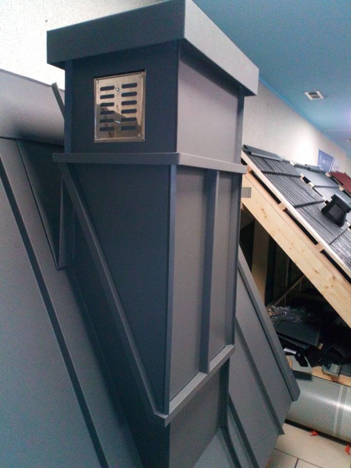 Szary komin wentylacyjny na dachu z blachy trapezowej, widoczne próbki dachówek w tle.