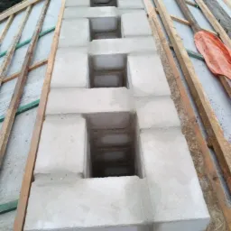 Wznoszony komin z bloczków betonowych na dachu, widoczne otwory wentylacyjne w konstrukcji, drewniane elementy więźby dachowej w tle.