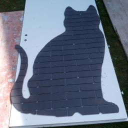 Damian Tychulski - Sylwetka kota ułożona z ciemnych płytek na białej płycie, leżącej na trawie, widoczne narzędzia i pozostałości materiałów budowlanych.