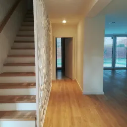 Nowoczesny korytarz z drewnianą podłogą, schodami z drewnianymi stopniami i ażurową białą ścianką działową, widok na otwarte drzwi i okna z żaluzjami.