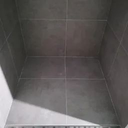 Szary, nowoczesny prysznic walk-in z odpływem liniowym i dużymi płytkami w jednolitym odcieniu szarości, bez brodzika.