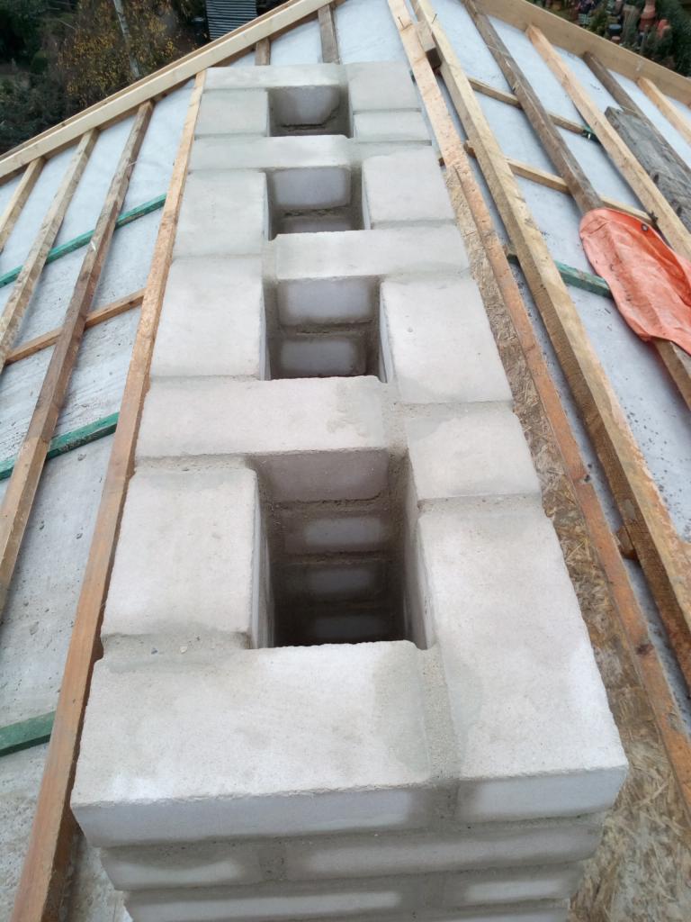 Wznoszony komin z bloczków betonowych na dachu, widoczne otwory wentylacyjne w konstrukcji, drewniane elementy więźby dachowej w tle.