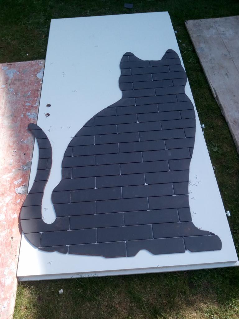 Sylwetka kota ułożona z ciemnych płytek na białej płycie, leżącej na trawie, widoczne narzędzia i pozostałości materiałów budowlanych.