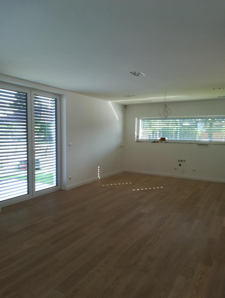 Jasne, puste wnętrze z drewnianą podłogą, dużymi oknami z żaluzjami i białymi ścianami, widoczne przygotowania do montażu oświetlenia sufitowego.