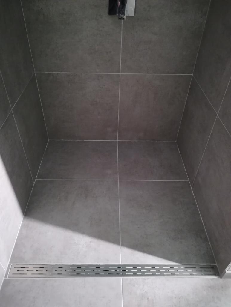 Szary, nowoczesny prysznic walk-in z odpływem liniowym i dużymi płytkami w jednolitym odcieniu szarości, bez brodzika.