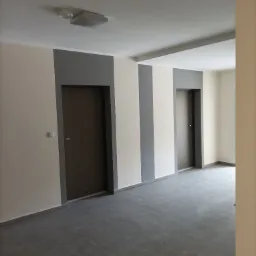 Wnętrze świeżo wykończonego pokoju z ciemną podłogą z płytek, dwoma brązowymi drzwiami z szarymi obramowaniami i ścianami w odcieniach bieli i szarości.