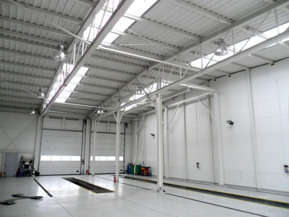 Wnętrze nowo wybudowanej hali z białymi ścianami, stalową konstrukcją dachu z oknami dachowymi, betonową posadzką z kanałami rewizyjnymi i zamkniętymi bramami segmentowymi.