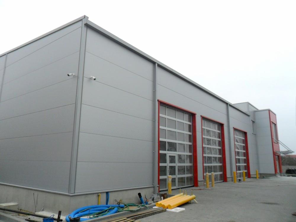 Zewnętrzny widok szarej hali z trzema segmentowymi bramami garażowymi z czerwonymi obramowaniami, widoczne rury i kable na zewnątrz budynku.