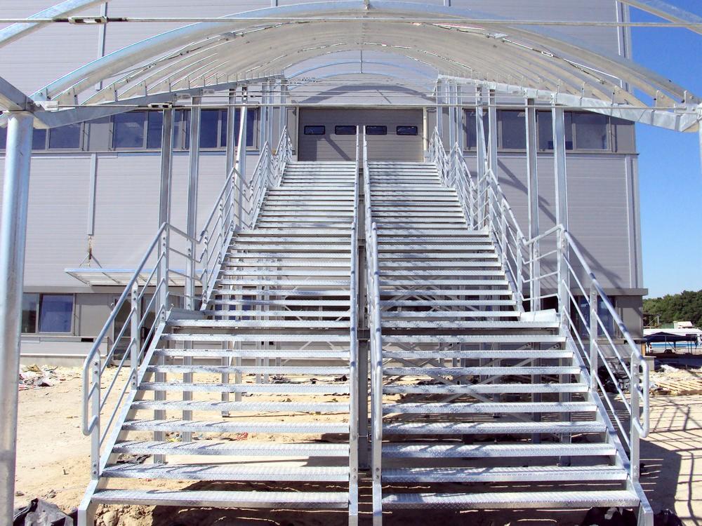 Metalowe schody zewnętrzne z zadaszeniem prowadzące do hali magazynowej, widoczne okna i elewacja z blachy trapezowej.