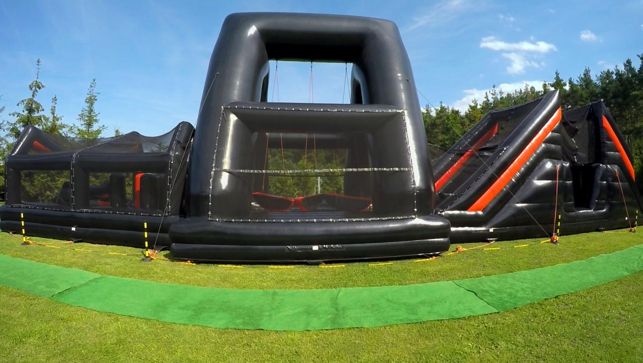 Czarny, dmuchany tor przeszkód z elementami do wspinania i huśtawką, rozstawiony na trawie w plenerze.