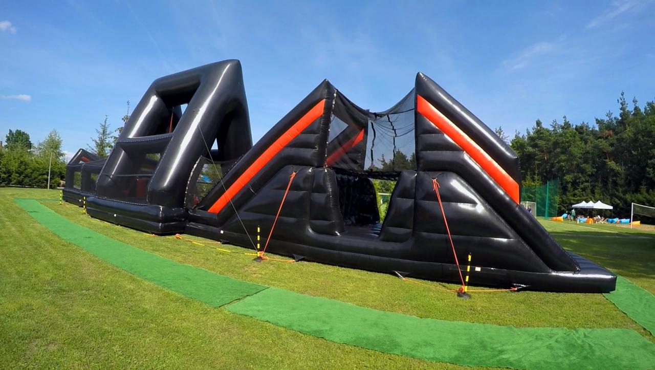 Długi, czarny, dmuchany tor przeszkód z pomarańczowymi akcentami, rozstawiony na trawiastym terenie podczas imprezy plenerowej, widoczne liny mocujące i zielone pasy podłoża.