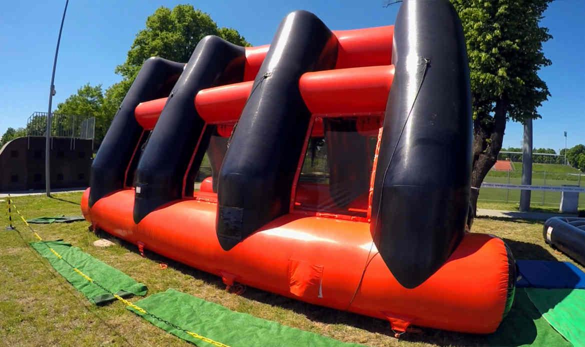 Pompowana konstrukcja przeszkód w kolorach pomarańczowym i czarnym, ustawiona na trawie, z widocznymi matami ochronnymi i taśmą zabezpieczającą teren.