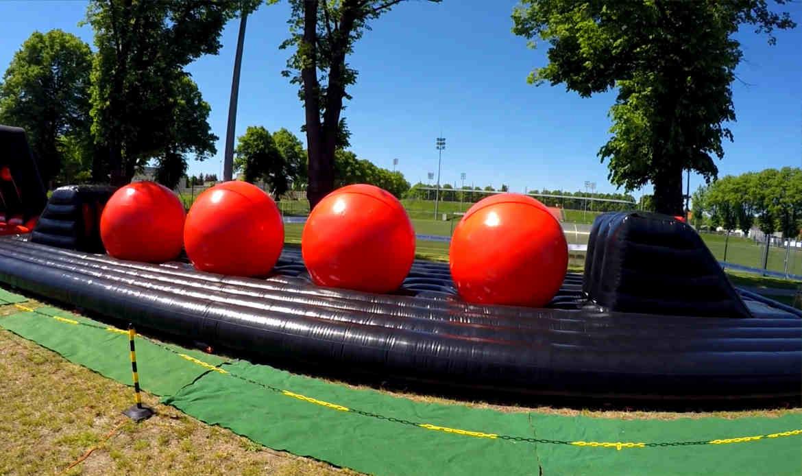 Nadmuchiwany tor przeszkód z czterema dużymi, czerwonymi kulami na czarnej, błyszczącej platformie, ustawiony na zielonej trawie w plenerze.