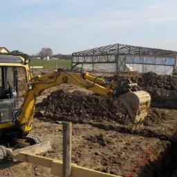 Żółta mini koparka wykonuje wykop pod fundamenty, widoczne drewniane szalunki, w tle budynki gospodarcze i dom jednorodzinny.