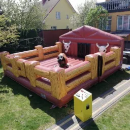 Nadmuchiwania atrakcja rodeo z elektrycznym panelem sterowania na trawie przed domem z żółtą elewacją.